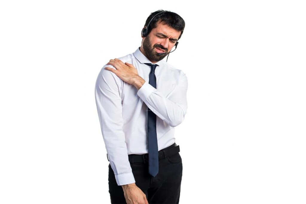 A man wearing a white shirt, black tie, and black pants is standing against a white background. Holding his left shoulder with a pained expression, he indicates discomfort or pain. He is also wearing a headset, possibly seeking physical therapy advice to alleviate his symptoms.