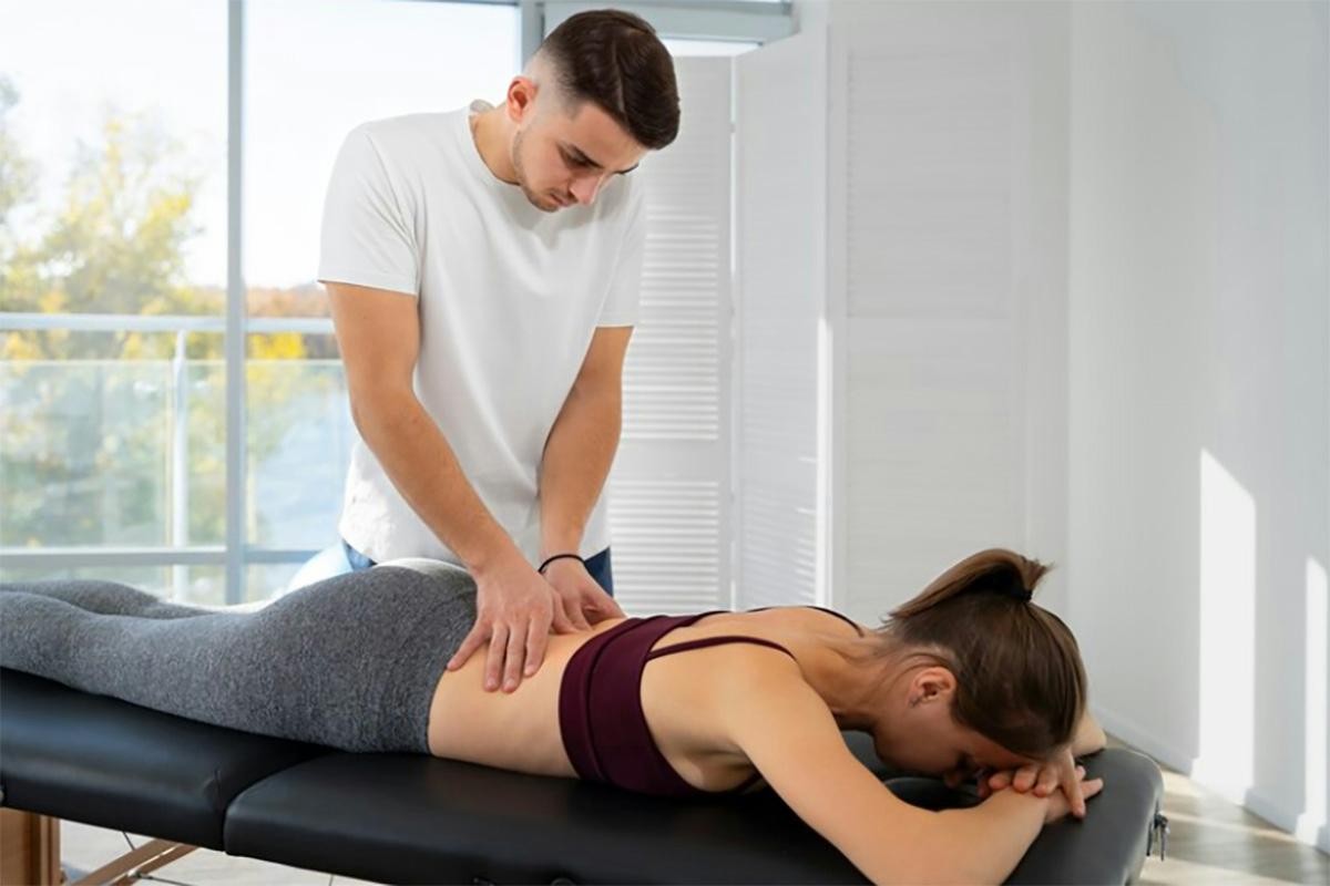 A person in a white shirt and short hair is giving a back massage, resembling physical therapy, to someone lying face down on a massage table. The person receiving the massage is wearing a maroon sports bra and gray pants. The setting is a bright room with large windows overlooking a scenic view.