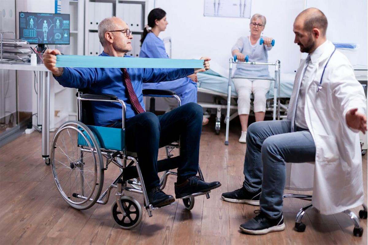 A man in a wheelchair exercises with a resistance band under the guidance of a doctor. In the background, a nurse observes another elderly woman standing with a walker. The room clearly serves as a physical therapy facility, focused on rehabilitation and recovery.
